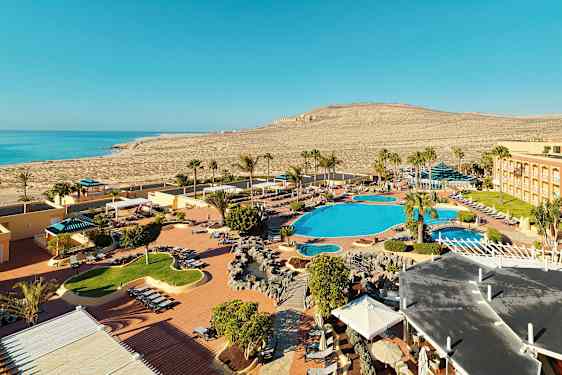 Bilden visar en semesteranläggning vid en kust med poolområden, solstolar och palmer. I bakgrunden syns havet och det omgivande torra landskapet. (AI-genererad text)