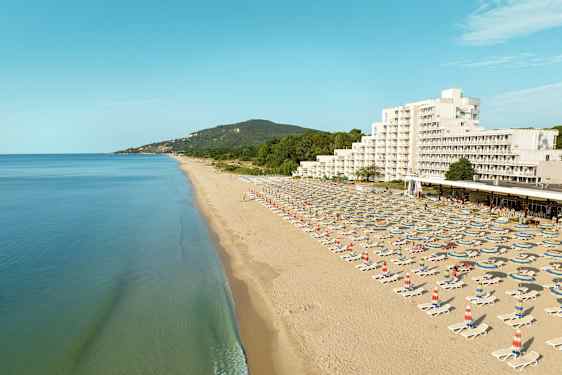 En lång sandstrand med solstolar och parasoller, samt ett hotell och gröna kullar i bakgrunden. (AI-genererad text)