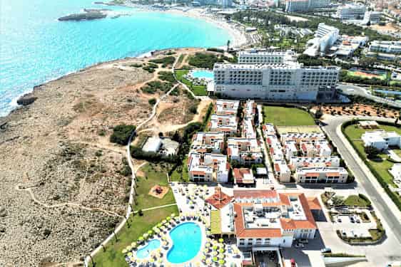 Flygbild över hotellkomplexet med pool, omgivande byggnader och en stenig kust med utsikt över havet. (AI-genererad text)