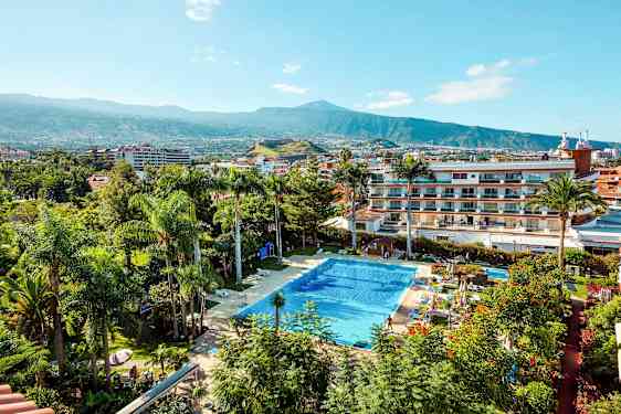 En pool omgiven av växter och byggnader med ett berg i bakgrunden. (AI-genererad text)