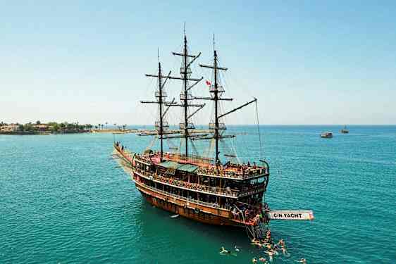 Ett stort träfartyg med segel som ligger i stilla vatten, omgiven av simmande personer, och i bakgrunden syns kustlinjen. (AI-genererad text)