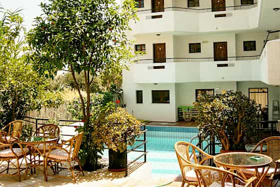 En utomhuspool omgiven av växtlighet och sittmöbler, samt en vit byggnad med balkonger i bakgrunden. (AI-genererad text)