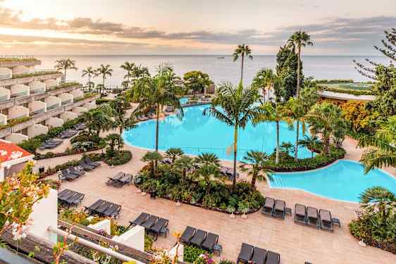 Bild på ett hotellområde med en stor swimmingpool omgiven av solstolar och tropiska växter. I bakgrunden syns havet och en solnedgång. (AI-genererad text)