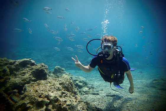 En person i dykardräkt under vatten omgiven av små fiskar och med en stenig havsbotten i bakgrunden. (AI-genererad text)