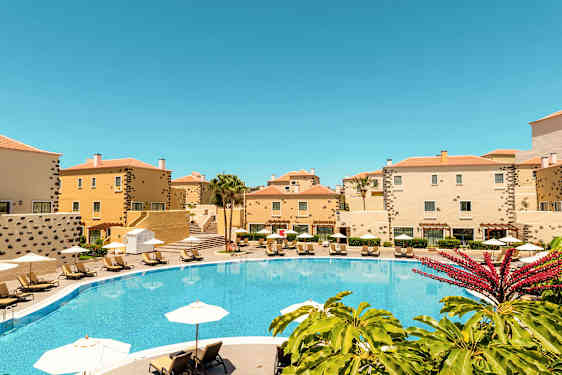 En pool omgiven av gula byggnader och solstolar under parasoller. (AI-genererad text)