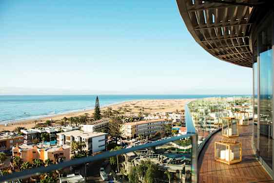 Utsikt över en strand med byggnader och en skog i förgrunden, sett från en balkong. (AI-genererad text)