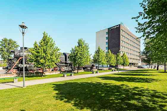 Ett gammalt ånglok står utomhus bredvid en modern hotellanläggning och grönskande träd. (AI-genererad text)