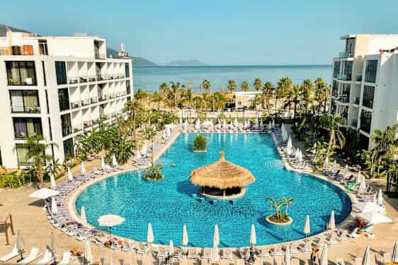 Hotellkomplex med stor pool omgiven av solstolar och parasoller, med havet i bakgrunden. (AI-genererad text)