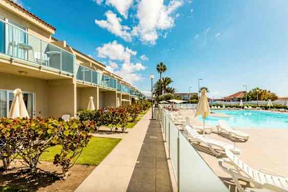 Bild av ett hotellområde med pool, solstolar och grönska. Byggnaden har balkonger. (AI-genererad text)