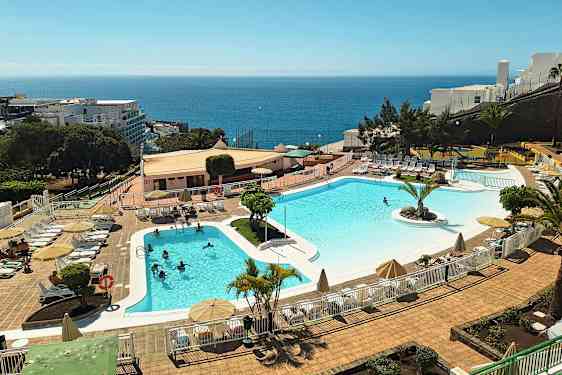 En utomhuspool vid ett semesterhotell med solstolar och parasoller. Havet syns i bakgrunden. (AI-genererad text)