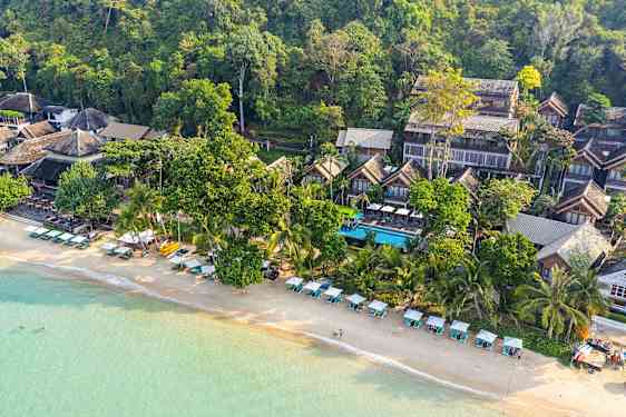 Flygfoto över en strand med solstolar och parasoller samt byggnader inbäddade i grönska. (AI-genererad text)