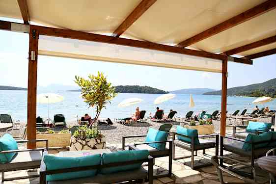 Utsikt från ett skuggigt sittområde mot en strand med solstolar och parasoller vid en lugn havsvik. (AI-genererad text)