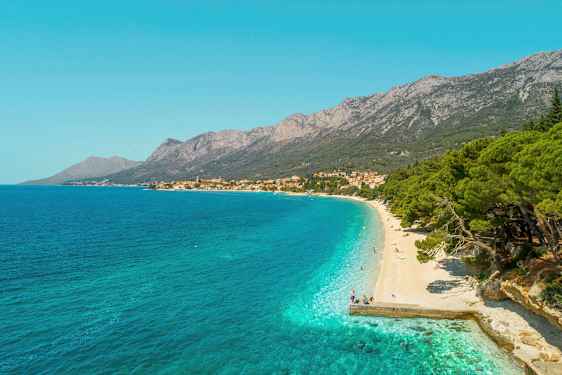 Vy över en klapperstensstrand med turkost vatten och grönskande träd. Berg och en by syns i bakgrunden. (AI-genererad text)