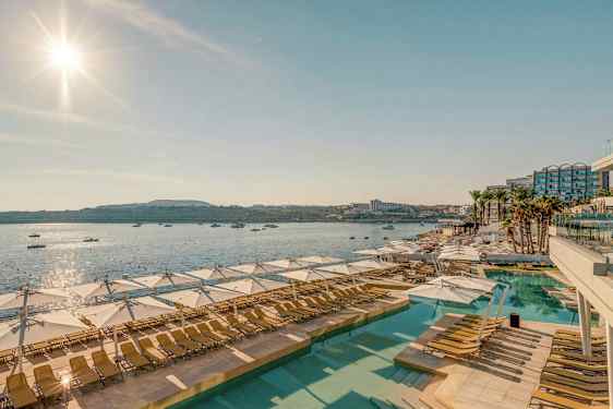Vy över en solig poolanläggning med solstolar och parasoller, med havet och båtar i bakgrunden. (AI-genererad text)