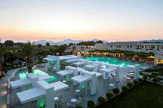 Modern hotellanläggning med pool och kvällsbelysning, omgiven av grönska och palmer med berg i bakgrunden. (AI-genererad text)