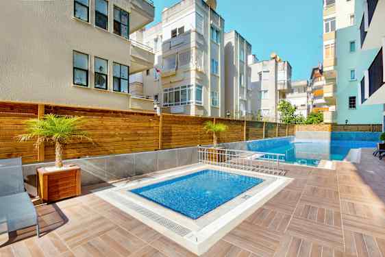 Utomhusområde med mindre pool och större bassäng omgivna av trädäck, palmer och närliggande byggnader. (AI-genererad text)