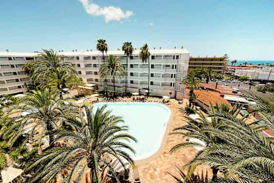 En utomhuspool omgiven av solstolar och palmer. I bakgrunden syns ett hotell med vit fasad. (AI-genererad text)