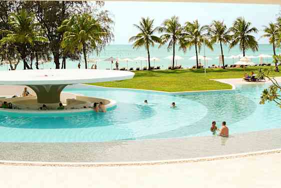 En pool med sittplatser i vattnet och en bar under ett stort tak. Bakom poolen finns gräsmatta, solstolar, parasoller, palmer och utsikt över havet. (AI-genererad text)