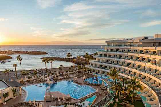 Hotellområde vid havet med utomhuspool och solstolar, omgiven av palmer, med en strand och vågbrytare i bakgrunden vid solnedgång. (AI-genererad text)