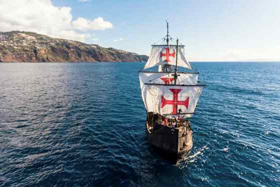 Ett segelfartyg med stora vita segel dekorerade med röda kors navigerar över en blå ocean i närheten av en kuperad kust. (AI-genererad text)