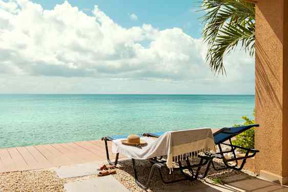 Två solstolar med en handduk och en hatt står bredvid ett lägre trädäck nära havet. En palmgren syns till höger. (AI-genererad text)