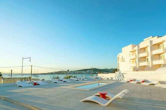 Utomhuspoolområde med moderna solstolar och utsikt mot havet samt vita byggnader i bakgrunden. (AI-genererad text)