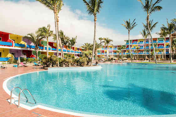 En stor pool omgiven av palmer, med färgglada hotellbyggnader i bakgrunden. (AI-genererad text)