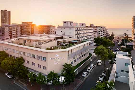 Hotellbyggnad vid havet omgiven av andra byggnader i solnedgång. (AI-genererad text)