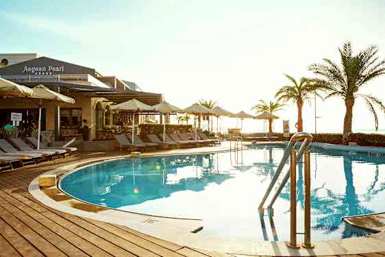 Utomhuspool med solstolar och parasoller kring. Byggnad med skylt 'Aegean Pearl' syns i bakgrunden. (AI-genererad text)
