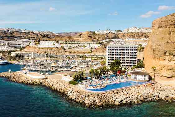 Bild av en kuststad med marina, byggnader på sluttning och hotell med pool. (AI-genererad text)