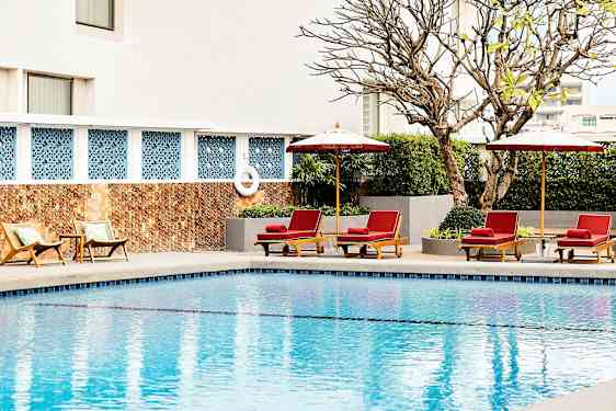 En pool med solstolar och parasoller vid kanten, omgivna av växter och en byggnadsvägg i bakgrunden. (AI-genererad text)