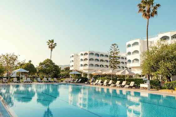 Hotellbyggnad med pool, solstolar och parasoller i förgrunden. (AI-genererad text)