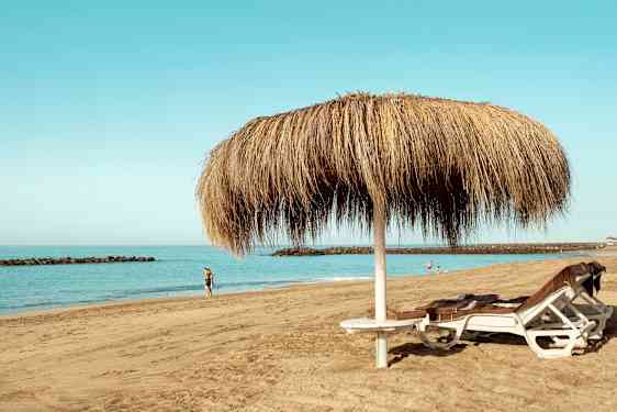 Bild av en sandstrand med två solstolar under ett parasoll av halm samt människor som går längs vattnet. (AI-genererad text)