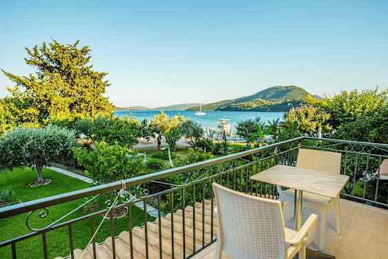 En balkong med bord och stolar, utsikt över en grön trädgård, havet och berg i bakgrunden. (AI-genererad text)
