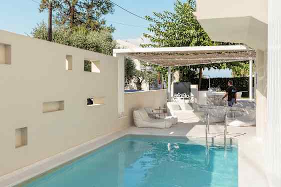 En pool omgiven av en ljus vägg med öppningar och en terrass med sittmöbler. (AI-genererad text)