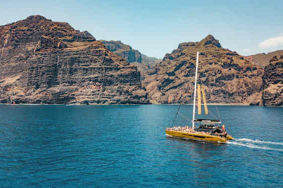 En katamaran seglar nära höga klippor, med klarblått vatten i förgrunden. (AI-genererad text)