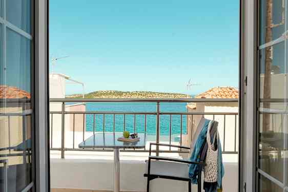 Utsikt från ett rum genom dörrar mot en balkong med bord, stol och klädesplagg, samt hav och landskap i bakgrunden. (AI-genererad text)