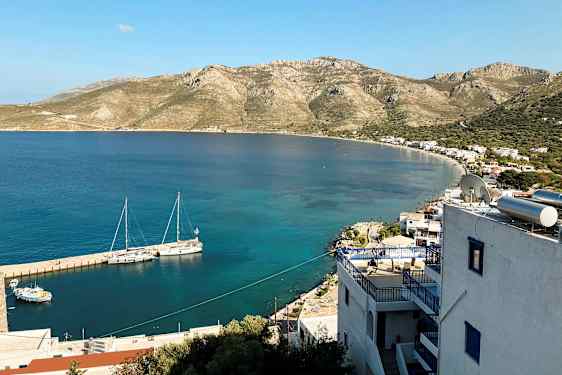 Vy över en kust med små byggnader och berg i bakgrunden. Båtar ligger förtöjda vid bryggan i det klara vattnet. (AI-genererad text)