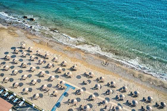 Flygfoto av en sandstrand med solstolar och parasoller, med kustlinjen och blått hav i bakgrunden. (AI-genererad text)