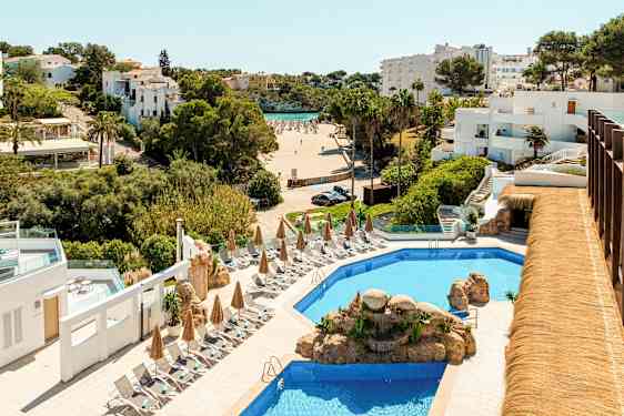 Hotellområde med en pool omgiven av solstolar och parasoller, i bakgrunden syns en strand och vitmålade byggnader. (AI-genererad text)