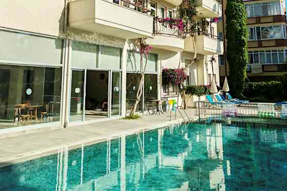 En pool framför en byggnad med balkonger och stora glasfönster. Bilderna visar även stolar och bord samt parasoller vid poolområdet. (AI-genererad text)