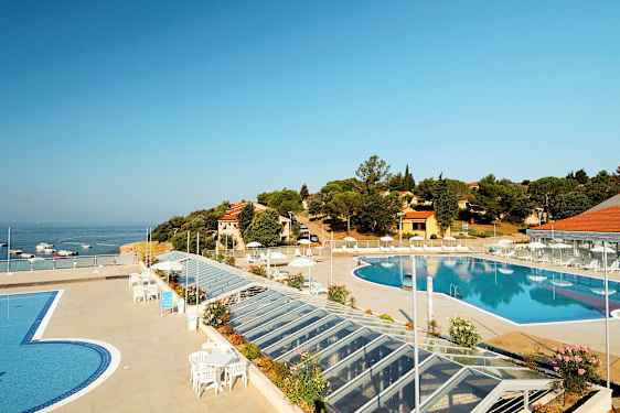 En vy över en utomhuspool omgiven av en terrass med solstolar och parasoller. I bakgrunden syns havet samt byggnader och vegetation. (AI-genererad text)