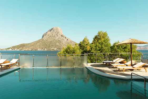 En utomhuspool med solstolar och parasoll, belägen nära havet med en kulle i bakgrunden. (AI-genererad text)