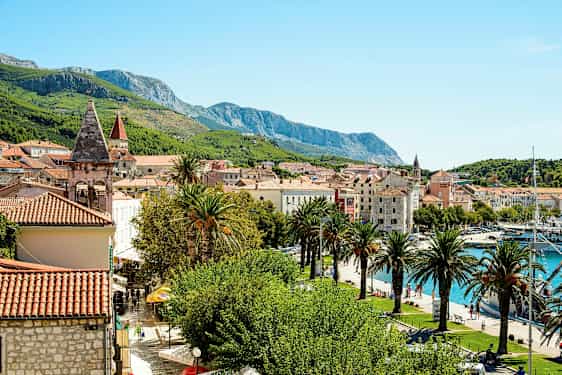 Bilden visar en pittoresk medelhavsstad med tegelröda tak, kyrktorn, palmer och gröna berg i bakgrunden. En småbåtshamn syns också vid vattnet. (AI-genererad text)