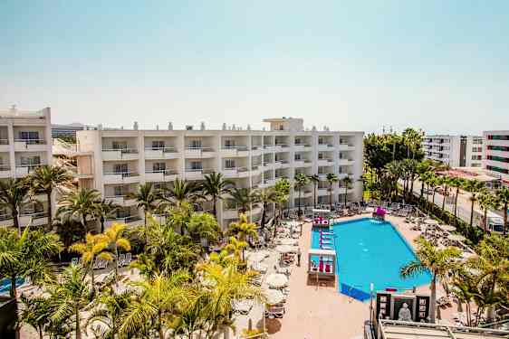 Hotellbyggnader med balkonger, en poolområde med solstolar och parasoller omgiven av palmer. (AI-genererad text)