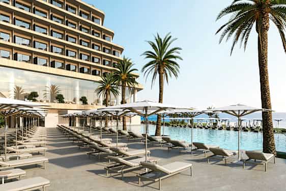 Bild föreställer en hotellpool med solstolar och parasoll i förgrunden, samt stora palmer och hotellfasaden i bakgrunden. (AI-genererad text)