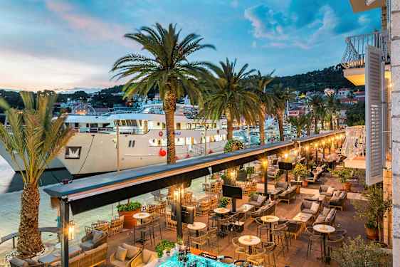 En vy av en utomhusrestaurang vid en hamn med palmer och stora båtar. (AI-genererad text)