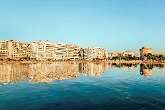 En stad vid vattnet med byggnader och ett runt torn reflekterade i det lugna vattnet. (AI-genererad text)