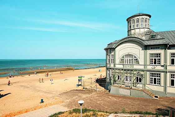 En strand med solbadande människor vid en lugn kustlinje, samt en större byggnad med klassisk arkitektur intill stranden. (AI-genererad text)