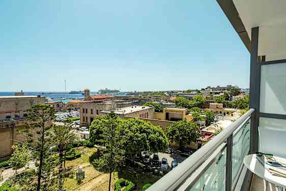 Utsikt från en balkong över en stad med historiska byggnader, träd och en hamn med båtar. (AI-genererad text)